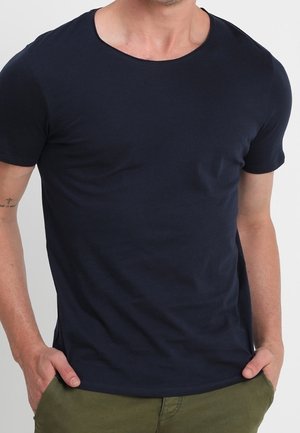 Mężczyzna ubrany w gładką czarną koszulkę z krótkim rękawem i okrągłym dekoltem, z rękami w kieszeniach oliwkowych spodni, widoczny tatuaż na lewej ręce.