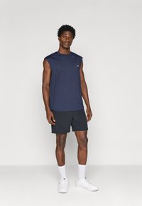 Jūrų pajėgų be rankovių sportinis marškinėlis, derinamas su juodomis šortais ir baltais sportbačiais. Marškinėliai turi apvalų iškirpimą ir subtilų logotipo detalę.