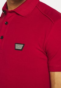 T-shirt polo rouge avec un col texturé, cinq boutons noirs et une écusson triangulaire en argent et noir sur la partie gauche de la poitrine.