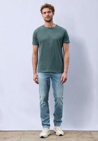T-shirt verde con colletto rotondo abbinato a jeans azzurri. Il modello è in piedi su una superficie piastrellata, indossando sneaker bianche, con uno sfondo semplice.