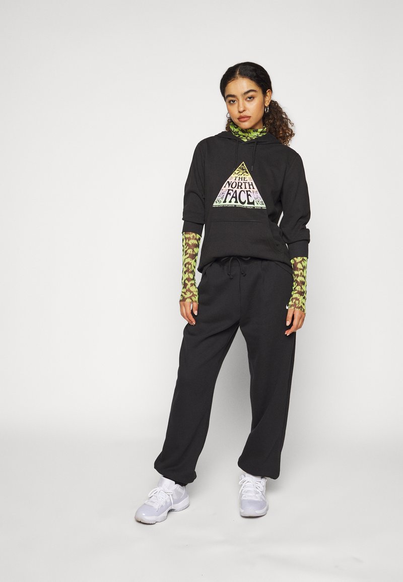 Schwarze, kürzere Kapuzenjacke mit einem dreieckigen Logo, kombiniert mit lockeren schwarzen Hosen und einem gemusterten grünen Langarmshirt. Weiße Sneaker runden den Look ab.
