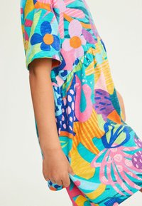 Värikäs kuviollinen mekko lyhyillä hihoilla, jossa on kukkia ja abstrakteja muotoja sinisessä, vaaleanpunaisessa, oranssissa ja vihreässä teal-taustalla.