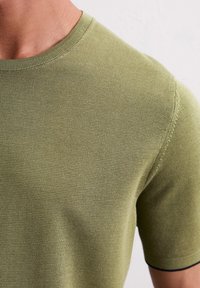 Oliwkowa, krótko rękawowa, dzianinowa koszulka z okrągłym dekoltem, z teksturowanym wzorem i ciemnoniebieskim akcentem wokół krawędzi rękawów.