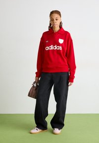 Felpa rossa con colletto polo, logo e emblema "adidas" bianchi. Pantaloni larghi neri, sneakers rosa e una piccola borsa marrone.