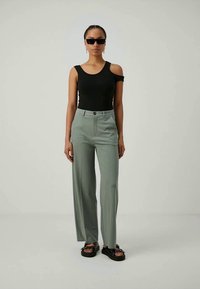 Mulher vestindo uma blusa assimétrica sem mangas preta, calças de cintura alta verde-claro, sandálias pretas de plataforma e óculos de sol retangulares pretos.