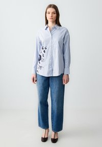 Camicia a righe azzurro chiaro con stampa floreale scura sul lato sinistro, abbinata a jeans dritti in denim blu a vita alta.