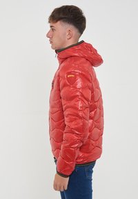 Giacca puffy rossa con finitura lucida, dotata di cappuccio, motivo trapuntato a zigzag e patch giallo sul spalla sinistra.