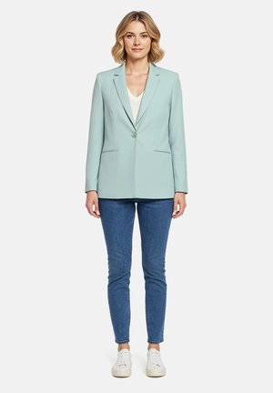 Femme aux cheveux blonds courts portant un blazer bleu clair, un haut blanc, un jean skinny bleu et des baskets blanches, debout face à l'avant.