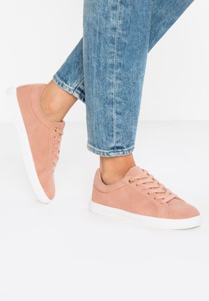 Personne portant un jean en denim bleu clair et des baskets roses avec des semelles blanches, debout sur une surface blanche.