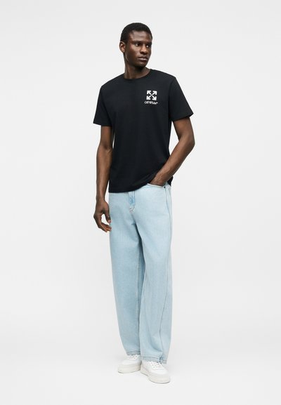 Άνδρας που φοράει μαύρο t-shirt με το λογότυπο Off-White, ανοιχτό μπλε παντελόνι με ευρύ μπατζάκι και άσπρα αθλητικά παπούτσια, στέκεται με το ένα χέρι στην τσέπη μπροστά σε λευκό φόντο.