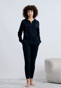 Svart zip-up huvtröja med röda detaljer, kombinerad med matchande joggers. Tillverkad av mjukt tyg, med avslappnad passform och framfickor.