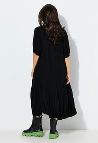 Vestido midi negro, fluido, con un diseño de capas y mangas cortas. Combinado con botas negras hasta el tobillo con suelas verdes. Textura de tela suave.