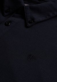 Mørk marineblå poloshirt med to sorte knapper på kraven og et subtilt broderet logo nedenfor slidsen.