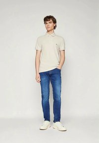 Jovem em pé com uma mão no bolso, vestindo uma t-shirt polo bege, jeans azuis e ténis brancos, contra um fundo claro e liso.
