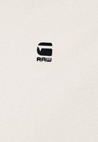 Beige bavlněné tričko s černým logem "RAW" uprostřed, s hladkou texturou a klasickým kulatým výstřihem.