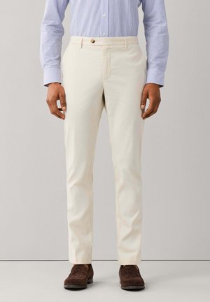 Man die crèmekleurige slim-fit broeken draagt, bruine suède schoenen en een blauw-wit gestreept overhemd met knopen, staand tegen een effen achtergrond.