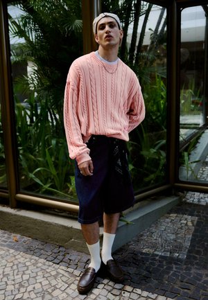 Fiatal férfi rózsaszín kábelmintás pulóverben, fekete rövidnadrágban, fehér zokniban, barna mokaszinban és hátul fordított bézs sapkában, egy csempézett úton áll.