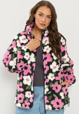 Jeune femme portant une veste pelucheuse à motif floral noir, blanc et rose sur un haut sombre et un jean bleu clair, posant avec une main près du col.