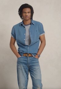 Farmer ing-shirt csíkos póló felett, világoskék farmernadrággal és barna övvel. Kiegészítők között óra és mandzsetta található.