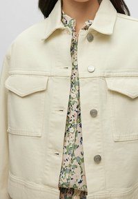 Krémová džínová bunda s dvěma náprsními kapsami, stříbrnými knoflíky a strukturovaným límcem. Pod ní je vidět blůza s květinovým vzorem.