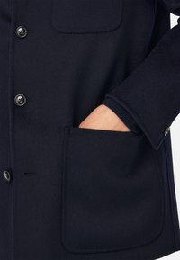 Tamsiai mėlynas vilnonis paltas su sagų uždarymu, krūtine kišene ir smulkintais rankogaliais. Audinys turi minkštą tekstūrą ir lygią apdailą.