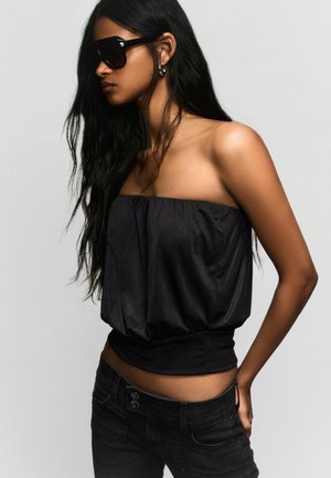 Kobieta o długich czarnych włosach, nosząca duże czarne okulary przeciwsłoneczne, czarny top bez ramiączek oraz czarne dżinsy na jednolitym tle.