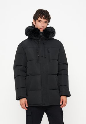 Homme portant une veste d’hiver matelassée noire avec une grande capuche noire bordée de fourrure, debout devant un fond clair uni.