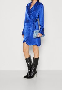 Modrá saténová zavinovací šaty s dlouhými rukávy, uvázaná v pase. Doplněná černými botami na vysokém podpatku s špičatou špičkou a malou modrou kabelkou ozdobenou flitry.