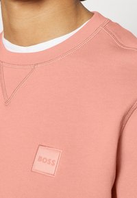 Růžová mikina s kulatým výstřihem vyrobená z příjemné látky, s obdélníkovou nášivkou s nápisem "BOSS" v kontrastní barvě a viditelnými detaily šití.