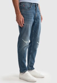Jeans de mezclilla azul con un corte relajado, que cuentan con dos roturas en las rodillas y un bolsillo delantero con una pequeña etiqueta roja. Llevados con zapatillas blancas.