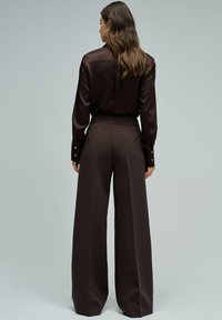 Mujer con cabello largo que lleva una blusa de satén marrón oscuro y pantalones de pierna ancha de tiro alto, de pie contra un fondo gris liso, de espaldas a la cámara.