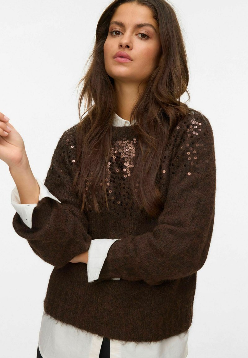 Femme aux longs cheveux bruns portant un pull marron foncé avec des sequins sur une chemise blanche, sur fond uni.