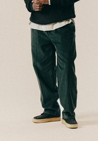 Σκούρα πράσινα παντελόνια καβούρι με φαρδιά εφαρμογή, συνδυασμένα με μαύρα παντόφλες που έχουν ανοιχτόχρωμη σόλα. Ορατές λεπτομέρειες υφής υφάσματος.