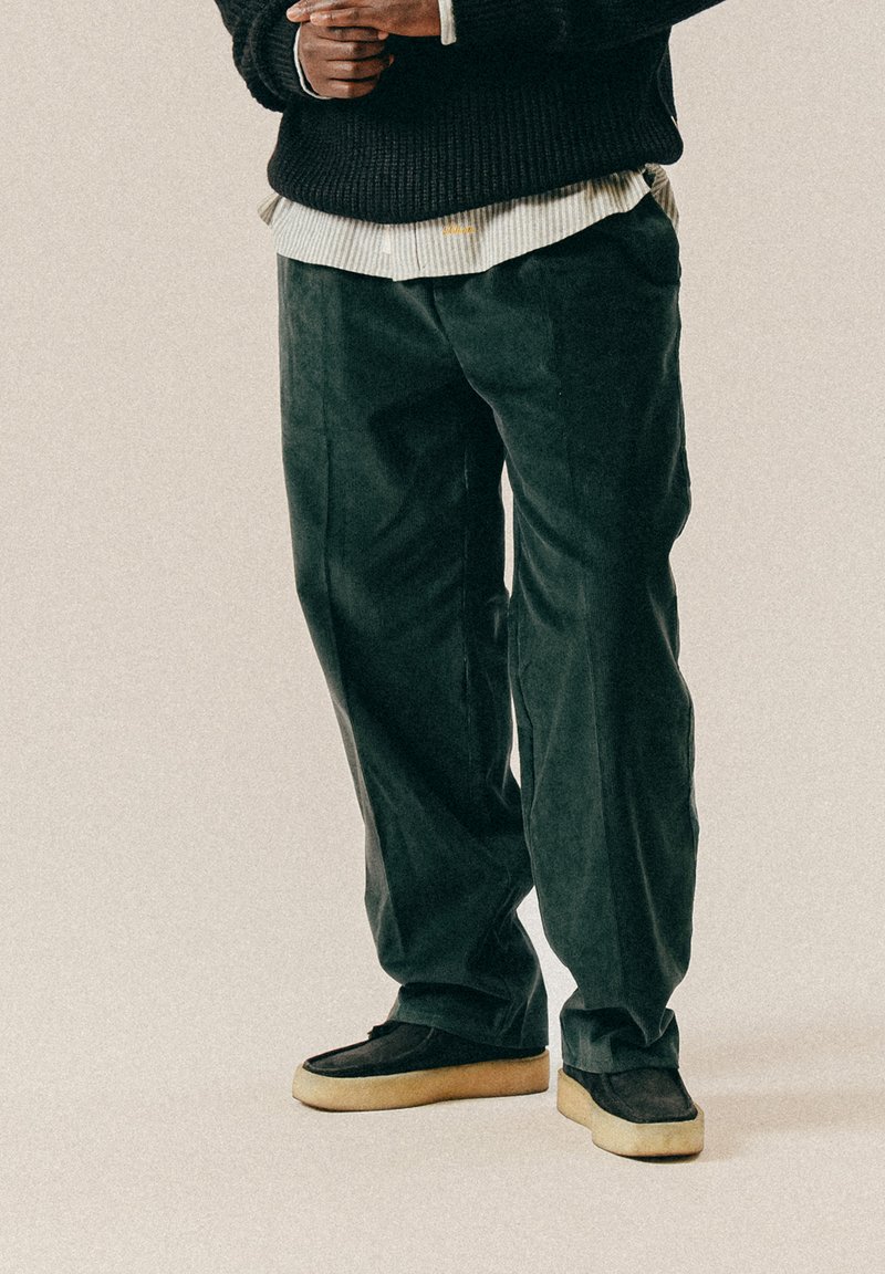 Σκούρα πράσινα παντελόνια καβούρι με φαρδιά εφαρμογή, συνδυασμένα με μαύρα παντόφλες που έχουν ανοιχτόχρωμη σόλα. Ορατές λεπτομέρειες υφής υφάσματος.