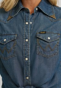 Denim shirt in medium blue, featuring snap buttons, decorative pockets with zigzag stitching, and a pointed collar with metallic accents.