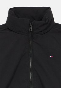 Schwarze Jacke mit hohem Kragen, Frontreißverschluss und kleinem rechteckigem Logo auf der Brust in den Farben Rot, Weiß und Blau.