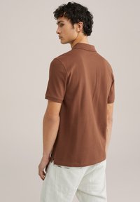 Brūns īsām piedurknēm polo krekls, izgatavots no kokvilnas, ar klasisku apkakli un sānu šķēlumiem, kopā ar gaišām biksēm.