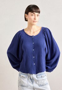 Navy blue textured blouse with a round neckline, button front, and voluminous, gathered sleeves. Paired with light denim shorts.