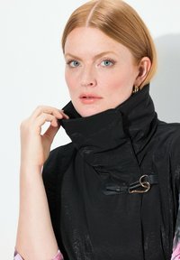 Schwarzer hochgeschlossener Mantel mit glänzenden Texturen und einem verstellbaren goldenen Verschluss. Glattes Material mit einer eleganten Silhouette.