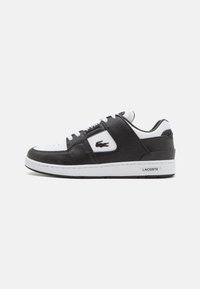 Sneaker bianco e nero con tomaia in pelle textrurata, lacci piatti e suola in gomma. Presenta un logo Lacoste sul lato.