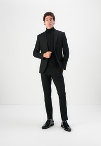 Fekete, slim fit szabású zakó, kifordított hajtókával, amelyet fekete garbóval párosítanak. A fényes fekete cipők teszik teljessé a megjelenést. Minimalista háttér.