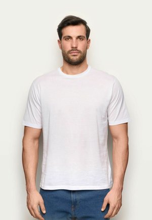 Uomo con capelli scuri e barba che indossa una semplice maglietta bianca e jeans blu, in piedi contro uno sfondo chiaro, rivolto in avanti.