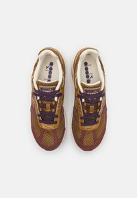 Zapatillas deportivas marrones y moradas con tela de malla, punta redonda, diseño de cordones, cuello acolchado y suela de goma contrastante.