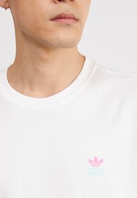 Balta medvilninė marškinėlių su apvaliu kaklu, su mažuoju rožiniu ir mėlynu Adidas logotipu, po kuriuo spausdinta „1972“. Trumpomis rankovėmis, glotnus paviršius.