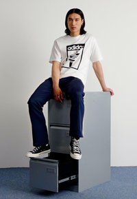 Gray metal filing cabinet with open drawer; topped by a person in a white graphic t-shirt and navy pants, seated casually. Black sneakers visible.