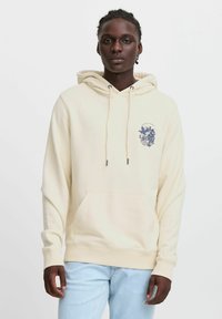 Haut à capuche couleur crème en tissu doux, avec une poche avant et un design graphique sur la zone gauche de la poitrine. Agrémenté de cordons de serrage.