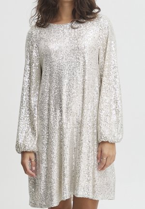 Femme portant une robe longue argentée à sequins, manches longues, col rond et coupe ample, debout devant un fond uni.