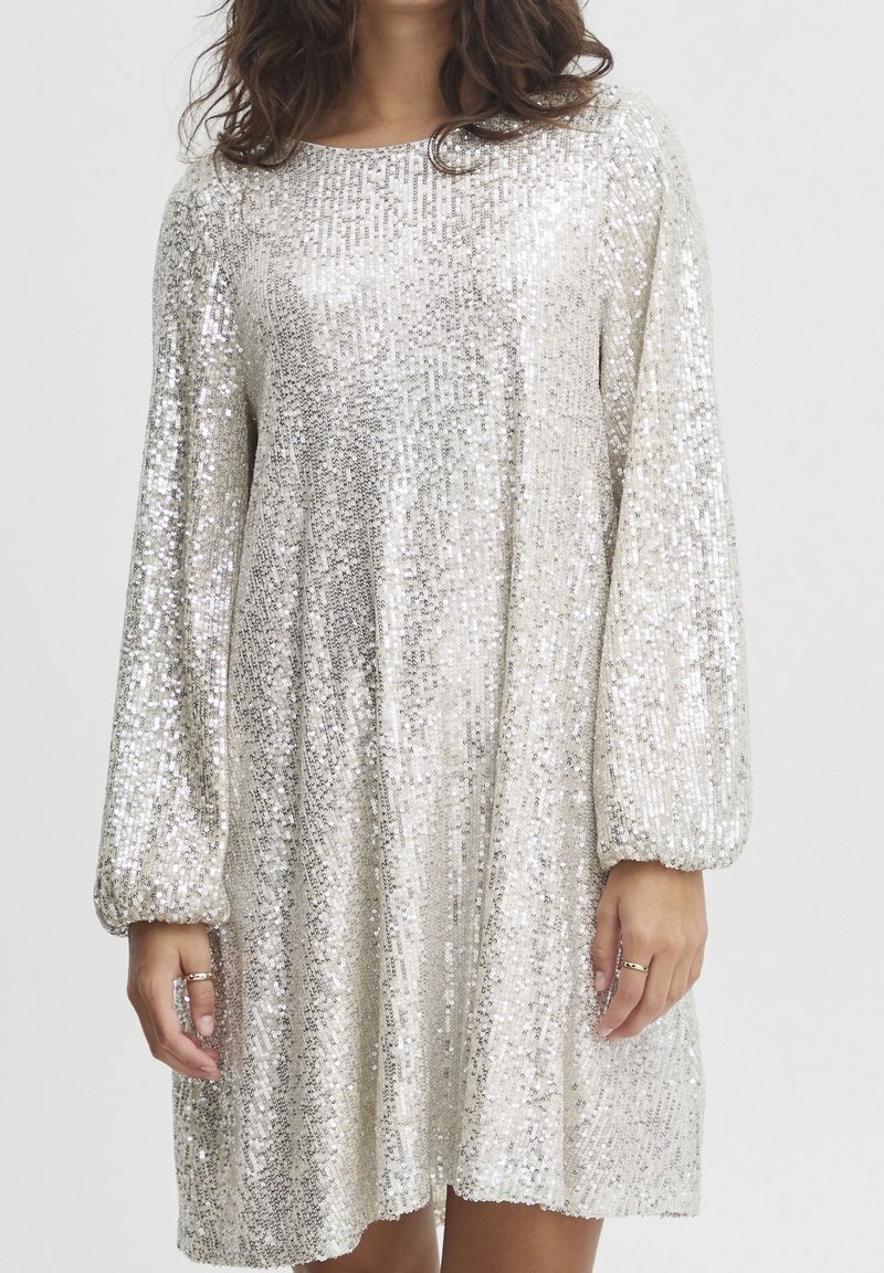 Femme portant une robe longue argentée à sequins, manches longues, col rond et coupe ample, debout devant un fond uni.