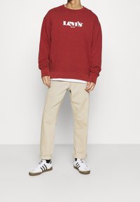 Felpa rossa con logo bianco "Levi's"; pantaloni beige; sneakers nere e bianche. Vestibilità casual con maniche lunghe e polsini a costine.