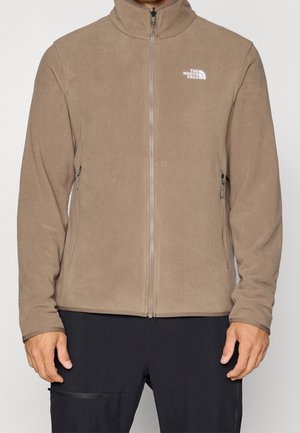 Taupefarbene Fleecejacke mit Reißverschluss, hohem Kragen, zwei seitlichen Reißverschlusstaschen und weißem "The North Face"-Logo auf der linken Brust, getragen mit schwarzer Hose.